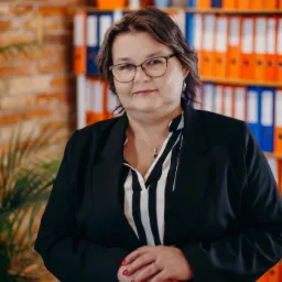 Portret kobiety w okularach i czarnym żakiecie na tle biura z regałem pełnym segregatorów w Gdańsku. Widoczne czerwone paznokcie i delikatna biżuteria.