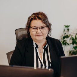 EUROMAX.PL SPÓŁKA Z OGRANICZONĄ ODPOWIEDZIALNOŚCIĄ SPÓŁKA KOMANDYTOWA - Portret kobiety w okularach i czarnej marynarce, siedzącej przy biurku z komputerem, w tle kalendarz i roślina doniczkowa.