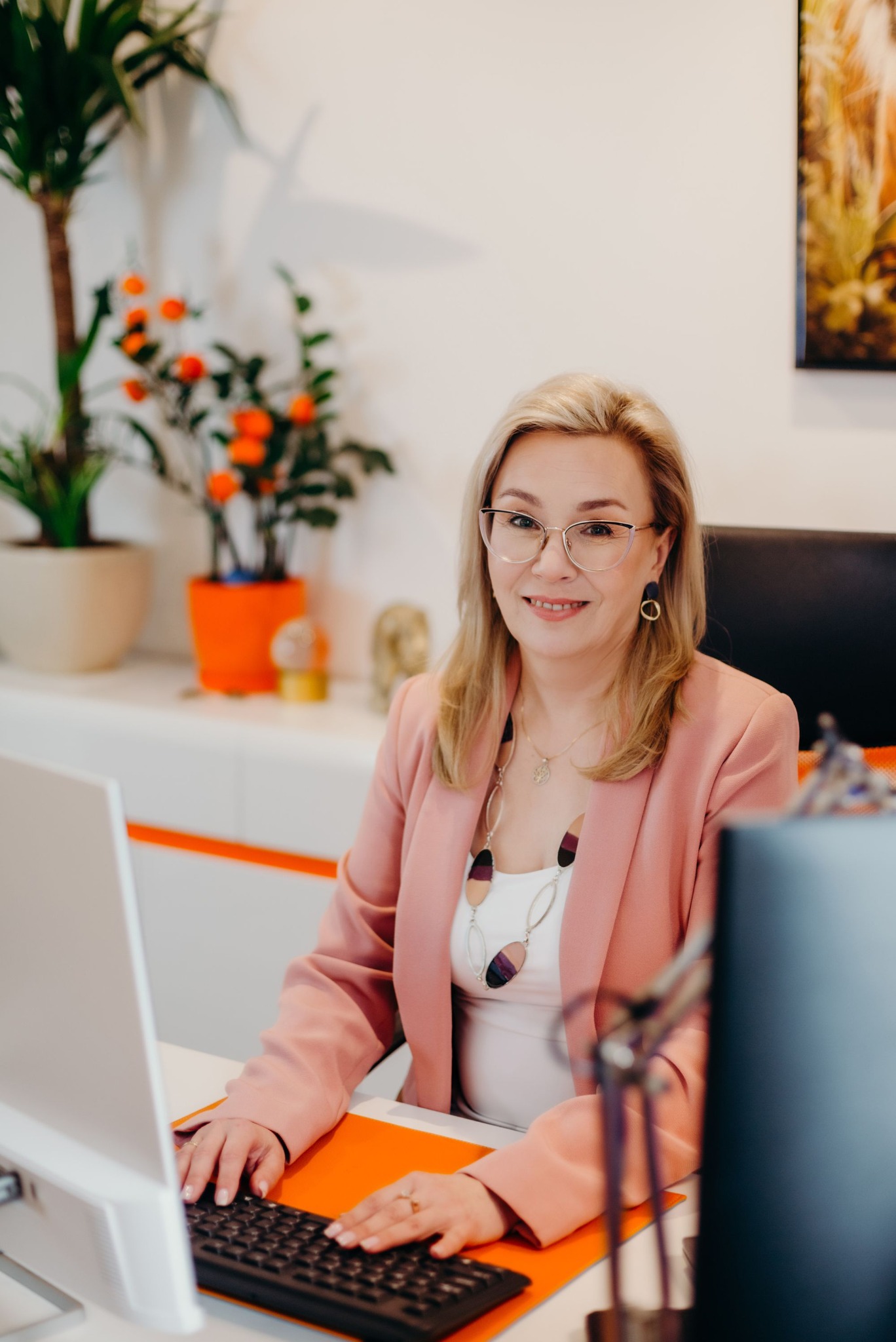 Uśmiechnięta blondynka w okularach i różowym żakiecie pracuje przy biurku z komputerem, na biurku pomarańczowa teczka.