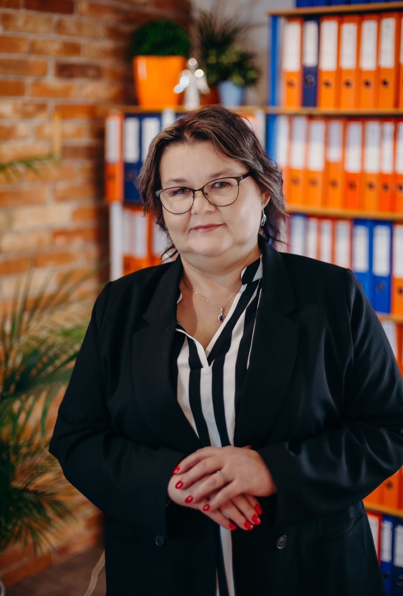 Portret kobiety w okularach i czarnym żakiecie na tle biura z regałem pełnym segregatorów w Gdańsku. Widoczne czerwone paznokcie i delikatna biżuteria.