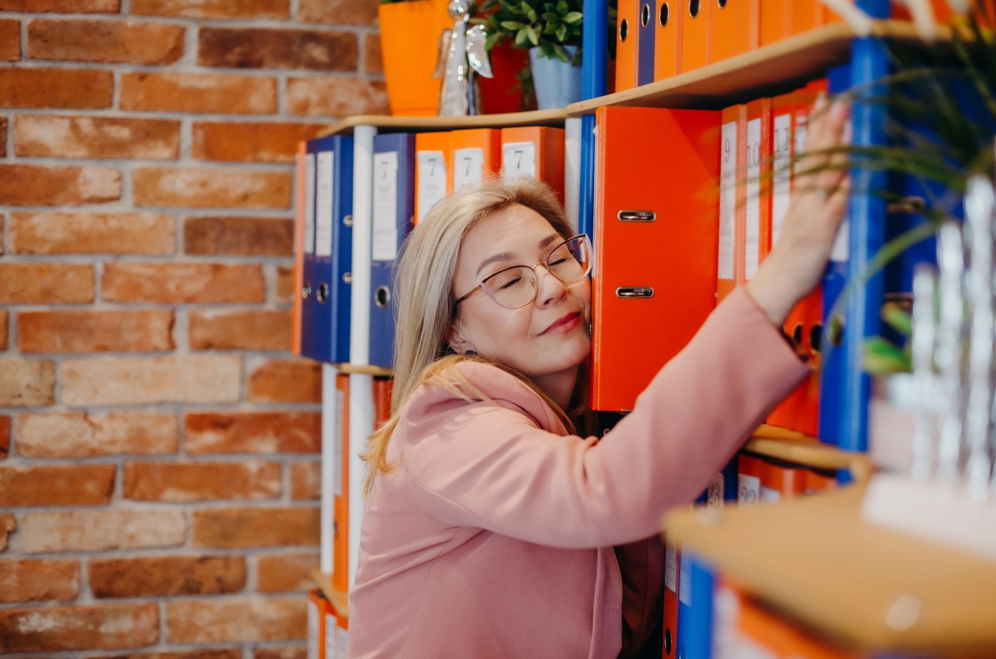 Uśmiechnięta kobieta w okularach przytula pomarańczowy segregator na półce z ceglanym tłem, obok niebieskie i pomarańczowe segregatory z numeracją.