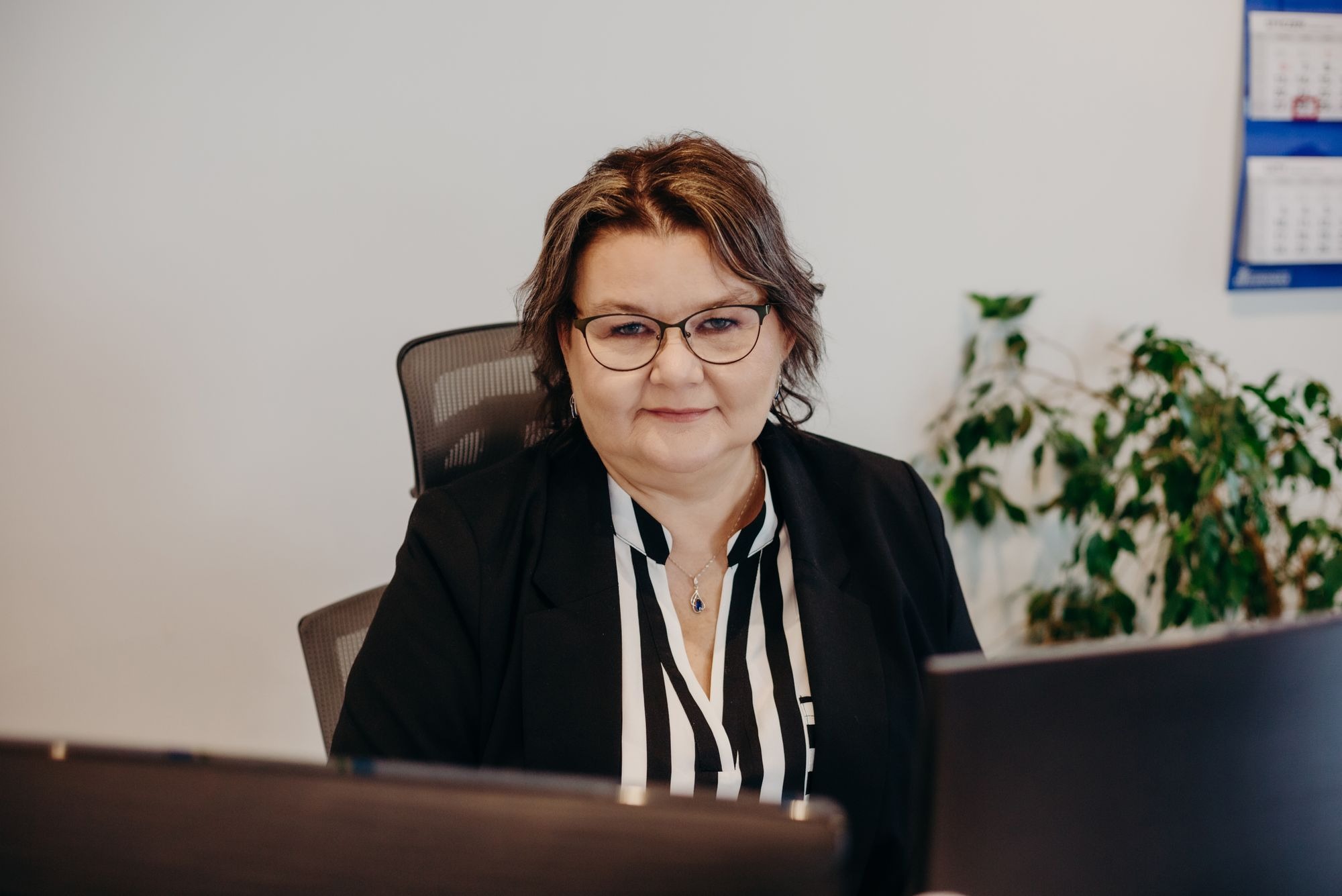 Portret kobiety w okularach i czarnej marynarce, siedzącej przy biurku z komputerem, w tle kalendarz i roślina doniczkowa.