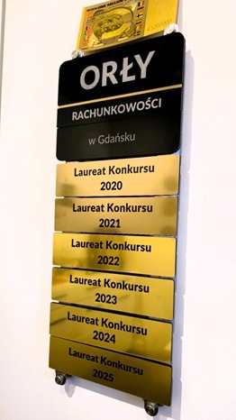 Tablica z nazwą 'Orły Rachunkowości w Gdańsku' i listą nagród 'Laureat Konkursu' z lat 2020-2025, umieszczona na białej ścianie. U góry złota ozdoba.