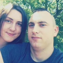 Portret uśmiechniętej kobiety z długimi brązowymi włosami i mężczyzny z krótkimi włosami na tle zieleni drzew.