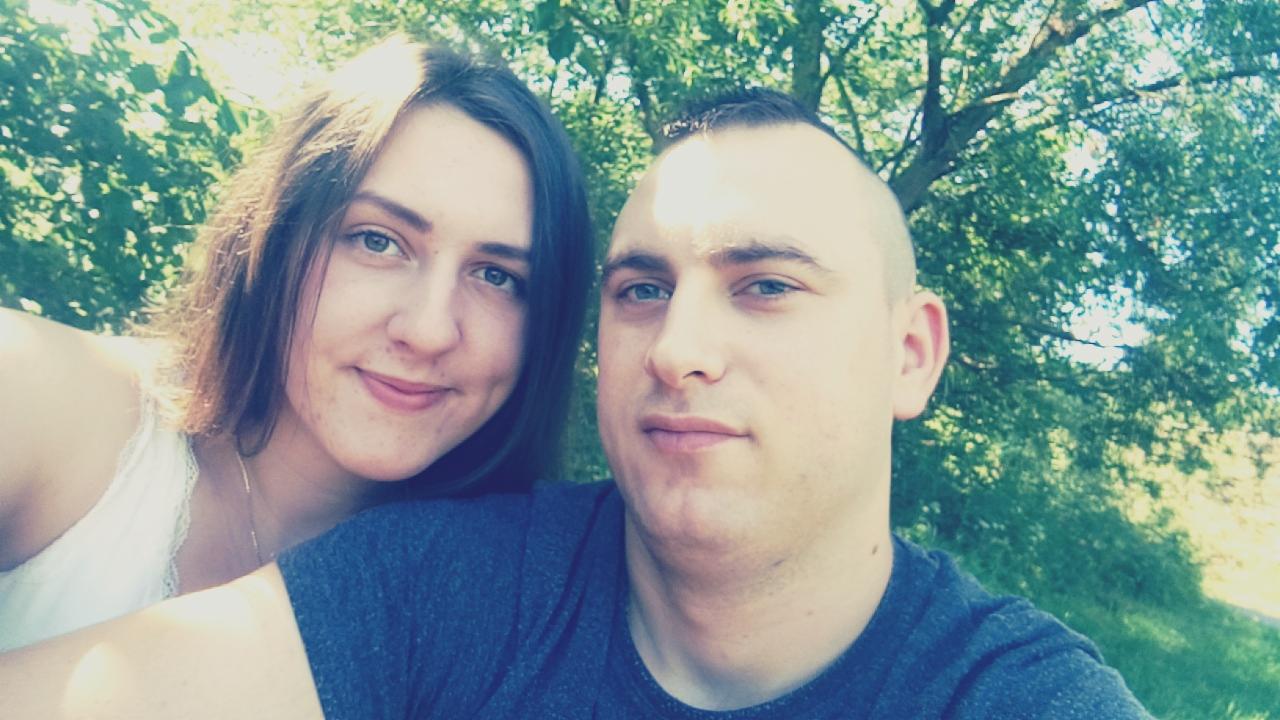 Portret uśmiechniętej kobiety z długimi brązowymi włosami i mężczyzny z krótkimi włosami na tle zieleni drzew.