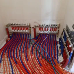 Rozdzielacze ogrzewania podłogowego z licznymi rurkami w kolorach czerwonym i niebieskim, ułożonymi na siatce grzewczej, widoczne notatki na ścianie.