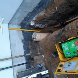 Wykop z widoczną rurą, folią kubełkową przy fundamencie, żółtą rurą gazową, skrzynką z zaworem gazowym, dwoma szpadlami i skrzynką z urządzeniem pomiarowym.