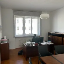 Wnętrze biura z dużym oknem z żaluzjami, biurkiem z komputerem i stosem dokumentów, oraz stołem konferencyjnym z czterema szklankami i długopisem. Na podłodze widoczny parkiet ułożony w jodełkę.