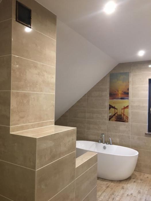 Nowoczesna łazienka na poddaszu z beżowymi płytkami imitującymi kamień, owalną białą wanną wolnostojącą i obrazem z motywem plaży na ścianie.