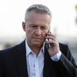 Mężczyzna w garniturze rozmawia przez telefon na tle nieokreślonego terenu.