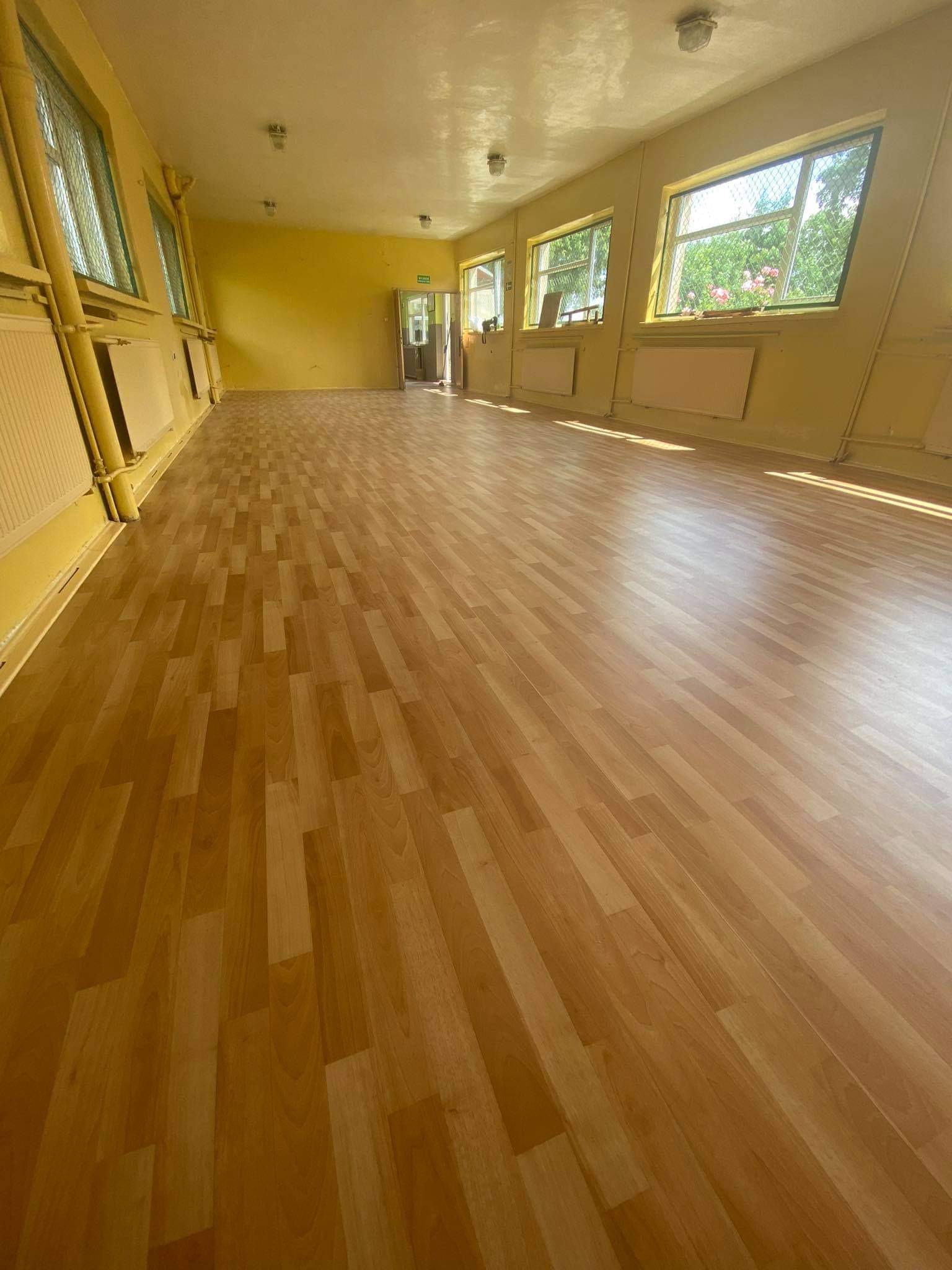 Jasna, przestronna sala z nową podłogą imitującą drewno, żółte ściany z grzejnikami pod oknami, widok na zieleń za oknem.