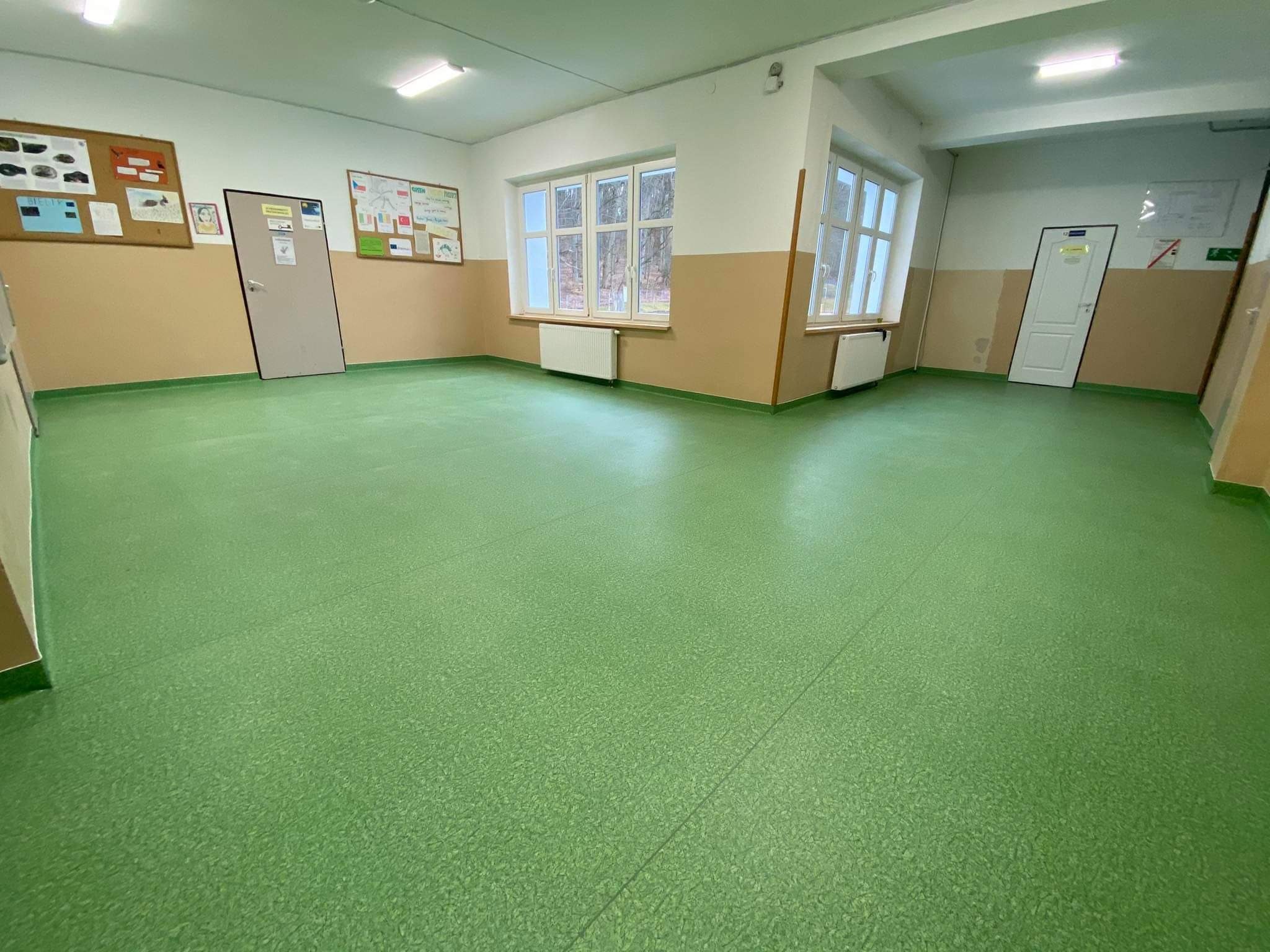 Jasnozielona, jednolita podłoga w przestronnym korytarzu szkolnym, ściany pomalowane na biało i beżowo, dwa okna z widokiem na drzewa, dwa grzejniki, dwoje drzwi oraz tablice informacyjne.