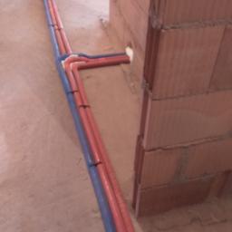 Fachmark - Wewnętrzne instalacje wodne z rur w izolacji, prowadzone po podłodze w surowym stanie, z widocznymi połączeniami i mocowaniami do podłoża.