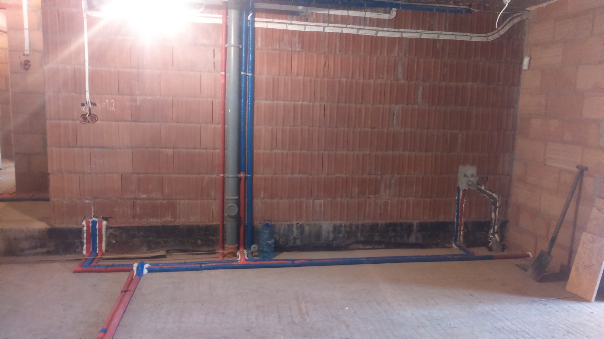 Surowy stan instalacji wodnej i kanalizacyjnej w budynku z czerwonej cegły, widoczne rury zasilające i odpływowe, w tle ściany z cegły i instalacja elektryczna pod sufitem.