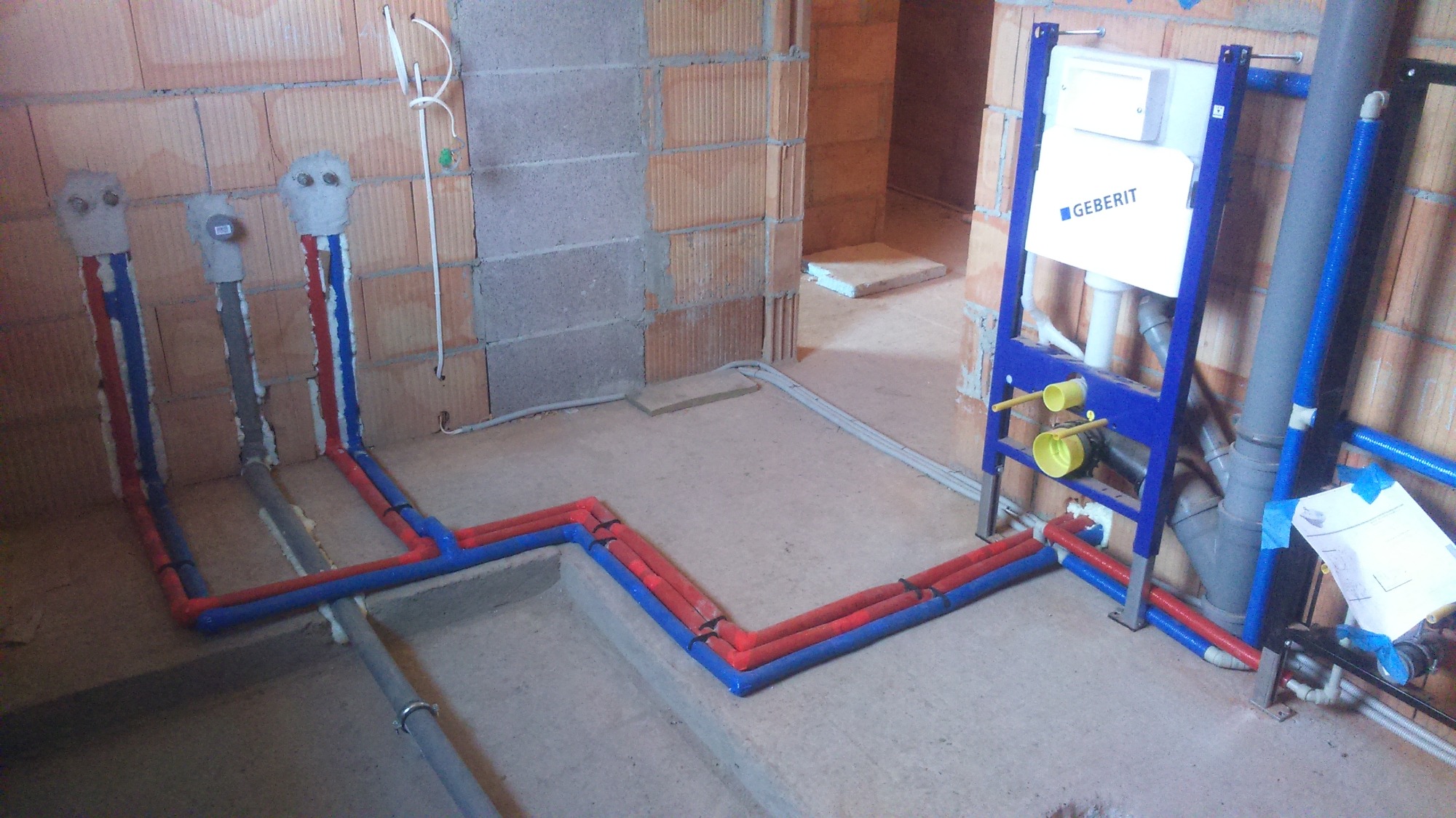 Surowe ściany z cegły dziurawki w łazience w trakcie instalacji hydraulicznej; widoczne rury ciepłej i zimnej wody w kolorze czerwonym i niebieskim, wystające ze ściany, oraz stelaż podtynkowy...