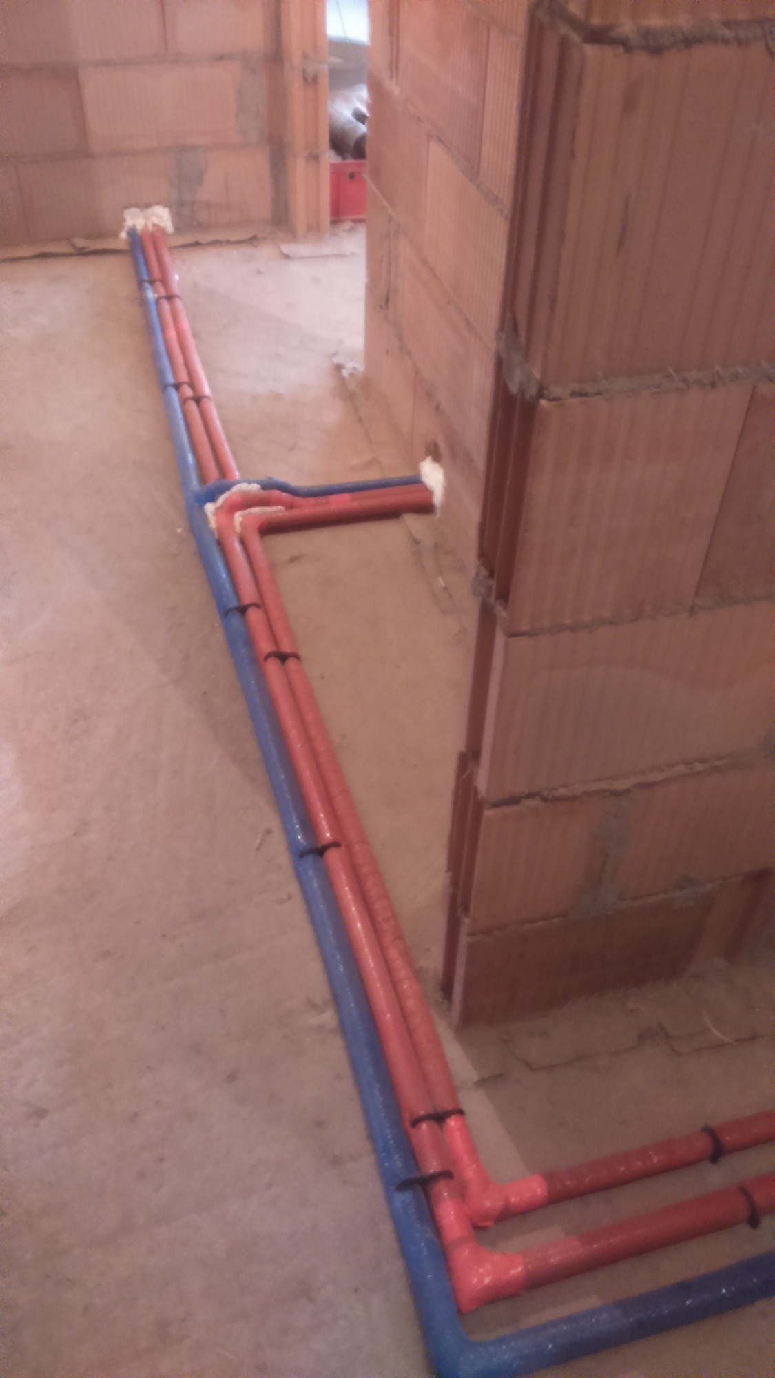 Wewnętrzne instalacje wodne z rur w izolacji, prowadzone po podłodze w surowym stanie, z widocznymi połączeniami i mocowaniami do podłoża.