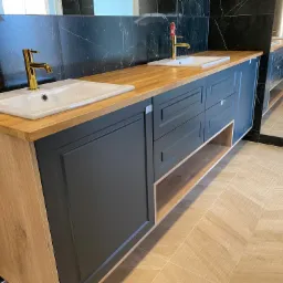 Nowoczesna łazienka z podwójną umywalką, drewnianym blatem i grafitowymi szafkami z frezowanymi frontami, złota armatura, czarne marmurowe płytki na ścianie i beżowa podłoga z płytek imitujących...