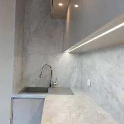 Nowoczesna kuchnia z blatem i ścianą imitującą marmur, srebrna bateria i szary zlew, oświetlenie LED pod szafkami.