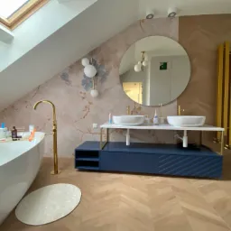 Nowoczesna łazienka na poddaszu z owalną wanną, umywalkami nablatowymi na szafce w kolorze granatowym i złotą armaturą.