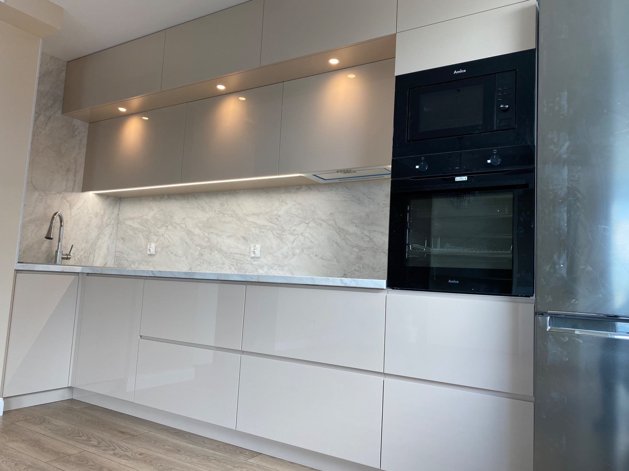 Jasna kuchnia z beżowymi, lakierowanymi frontami, marmurowym blatem i sprzętami marki Amica, oświetlona punktowo i listwą LED pod szafkami wiszącymi.