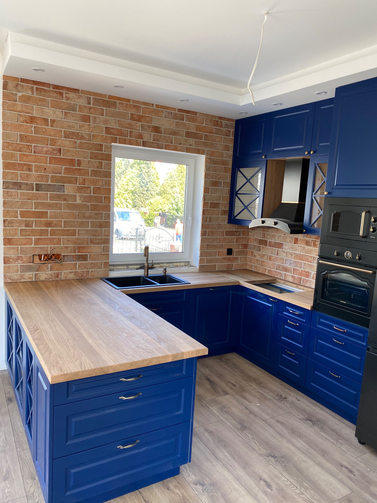 Niebieska kuchnia na wymiar z drewnianym blatem, ceglaną ścianą i widocznymi przewodami elektrycznymi w otworze, z czarnym piekarnikiem i okapem.