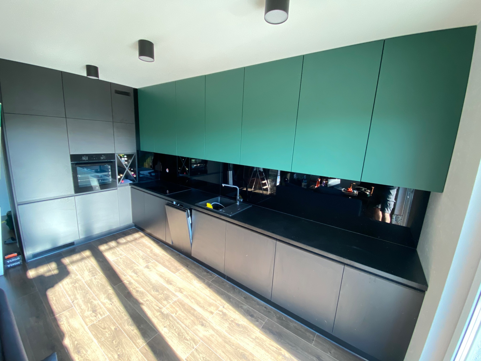 Nowoczesna kuchnia: ciemnoszare dolne szafki, butelkowa zieleń górnych szafek, czarny blat i lustrzany panel nad blatem, drewniana podłoga, oświetlenie sufitowe.