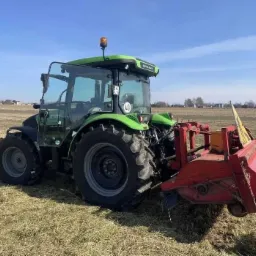 Zielony ciągnik Deutz-Fahr z czerwoną kosiarką rotacyjną na polu z resztkami skoszonej trawy, widoczne zabudowania w oddali pod błękitnym niebem.