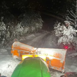 Widok z kabiny traktora na pomarańczowy pług odśnieżający drogę w nocy, oświetloną reflektorami, z zalesionym tłem pokrytym śniegiem i zwisającymi soplami lodu z góry.