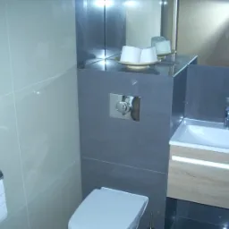 Nowoczesna łazienka z szarymi płytkami, podświetlaną szafką pod umywalką i minimalistycznym zestawem toaletowym. Widoczny chromowany przycisk spłuczki i uchwyt na papier toaletowy.