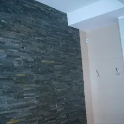 Ściana wyłożona ciemnym, łupkowym kamieniem dekoracyjnym ułożonym w poziome pasy, sąsiadująca z jasną ścianą z wieszakami na ubrania.