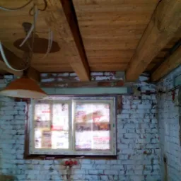 Surowy strop z drewnianych belek, okno zabezpieczone gazetami, ściany z cegły pomalowane na biało, widoczne instalacje elektryczne i lampa wisząca w trakcie remontu.