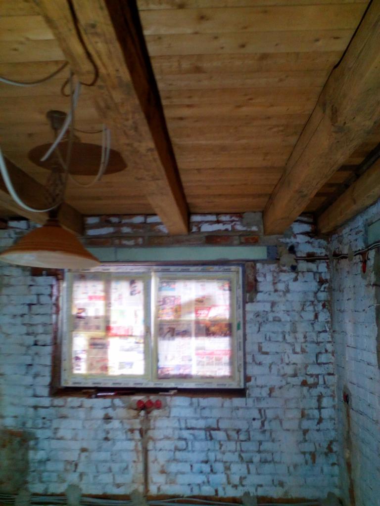 Surowy strop z drewnianych belek, okno zabezpieczone gazetami, ściany z cegły pomalowane na biało, widoczne instalacje elektryczne i lampa wisząca w trakcie remontu.