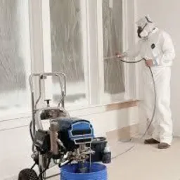 Pracownik w kombinezonie ochronnym maluje ramę okienną natryskowo, widoczny agregat malarski i zabezpieczone okno folią.