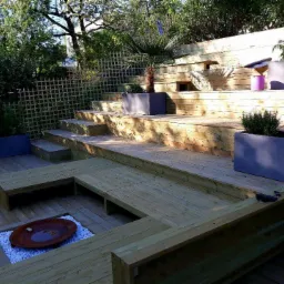 Nowoczesny taras drewniany z wielopoziomową konstrukcją, drewnianymi siedziskami wokół paleniska wypełnionego białymi kamieniami i roślinami w prostokątnych donicach.