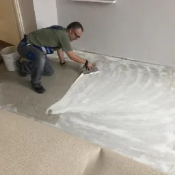 Mężczyzna w okularach rozprowadza klej szpachelką na podłodze przygotowując ją pod montaż beżowej wykładziny dywanowej.