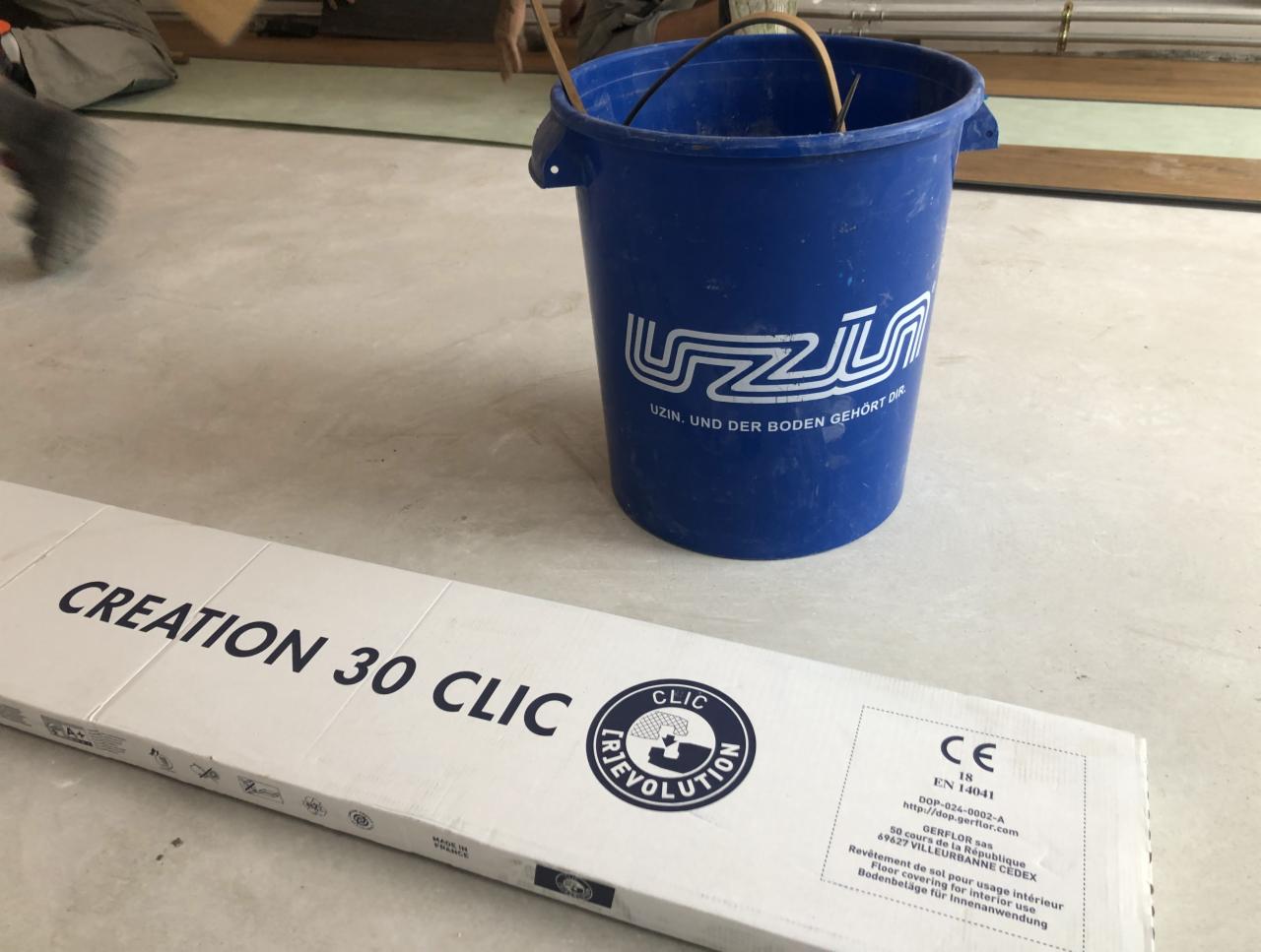 Montaż paneli podłogowych Creation 30 Clic: widoczny fragment podłogi w trakcie układania, paczka paneli z logo Gerflor oraz niebieskie wiadro Uzin