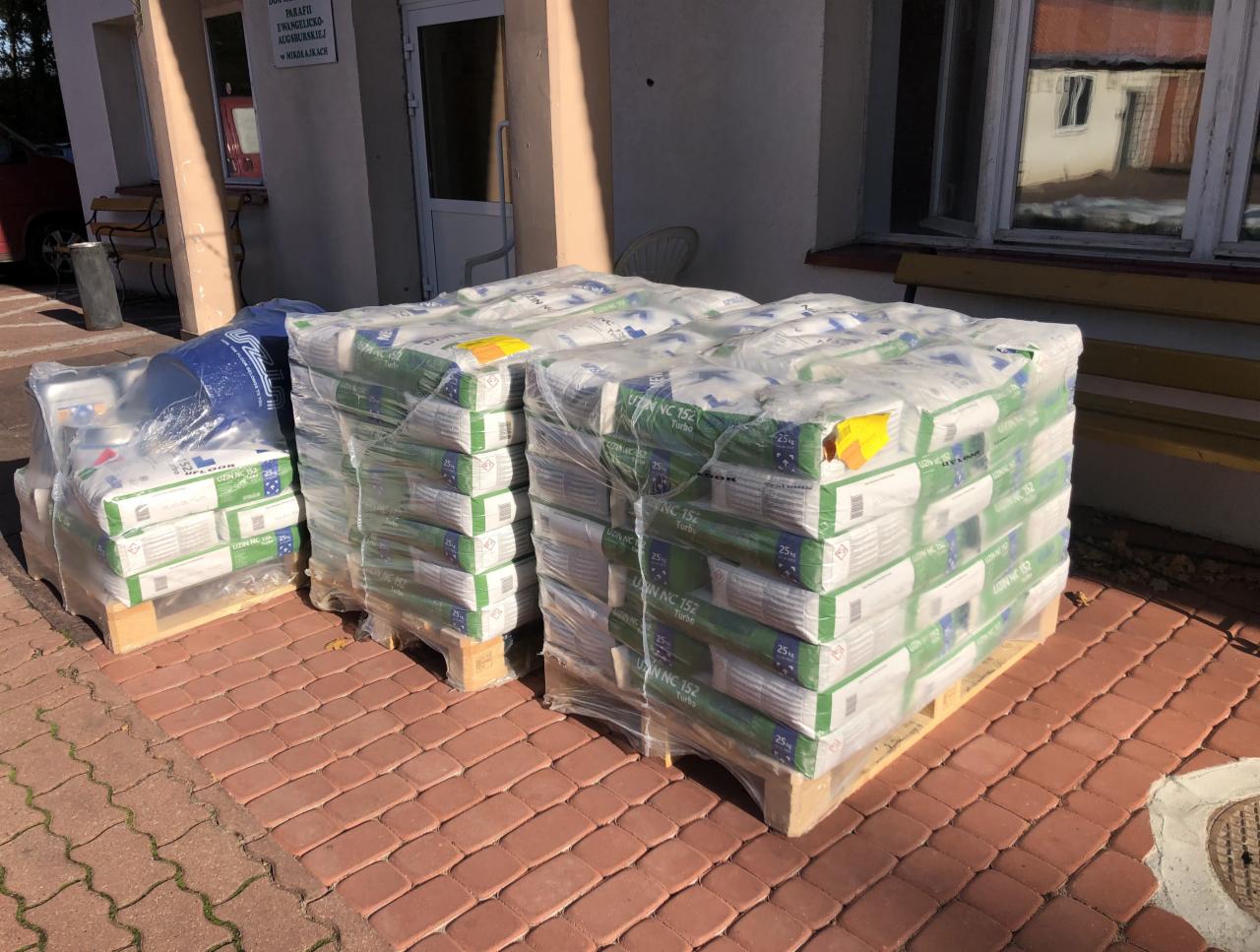 Dwa palety z workami kleju UZIN NC 152 Turbo, zapakowane w folię, ustawione na czerwonej kostce brukowej przed budynkiem z białymi drzwiami i oknem.