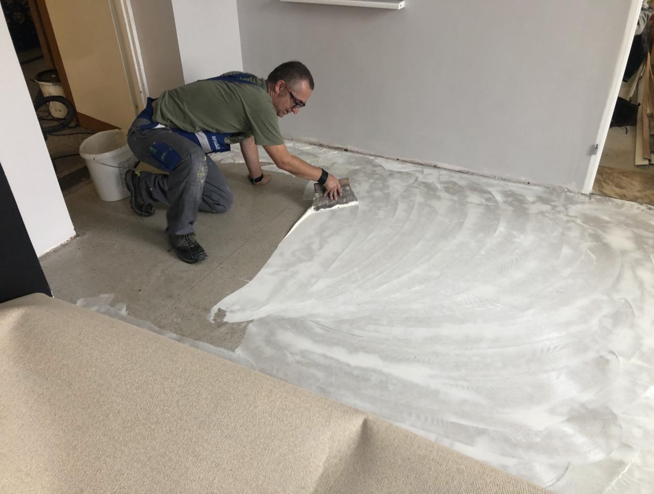 Mężczyzna w okularach rozprowadza klej szpachelką na podłodze przygotowując ją pod montaż beżowej wykładziny dywanowej.