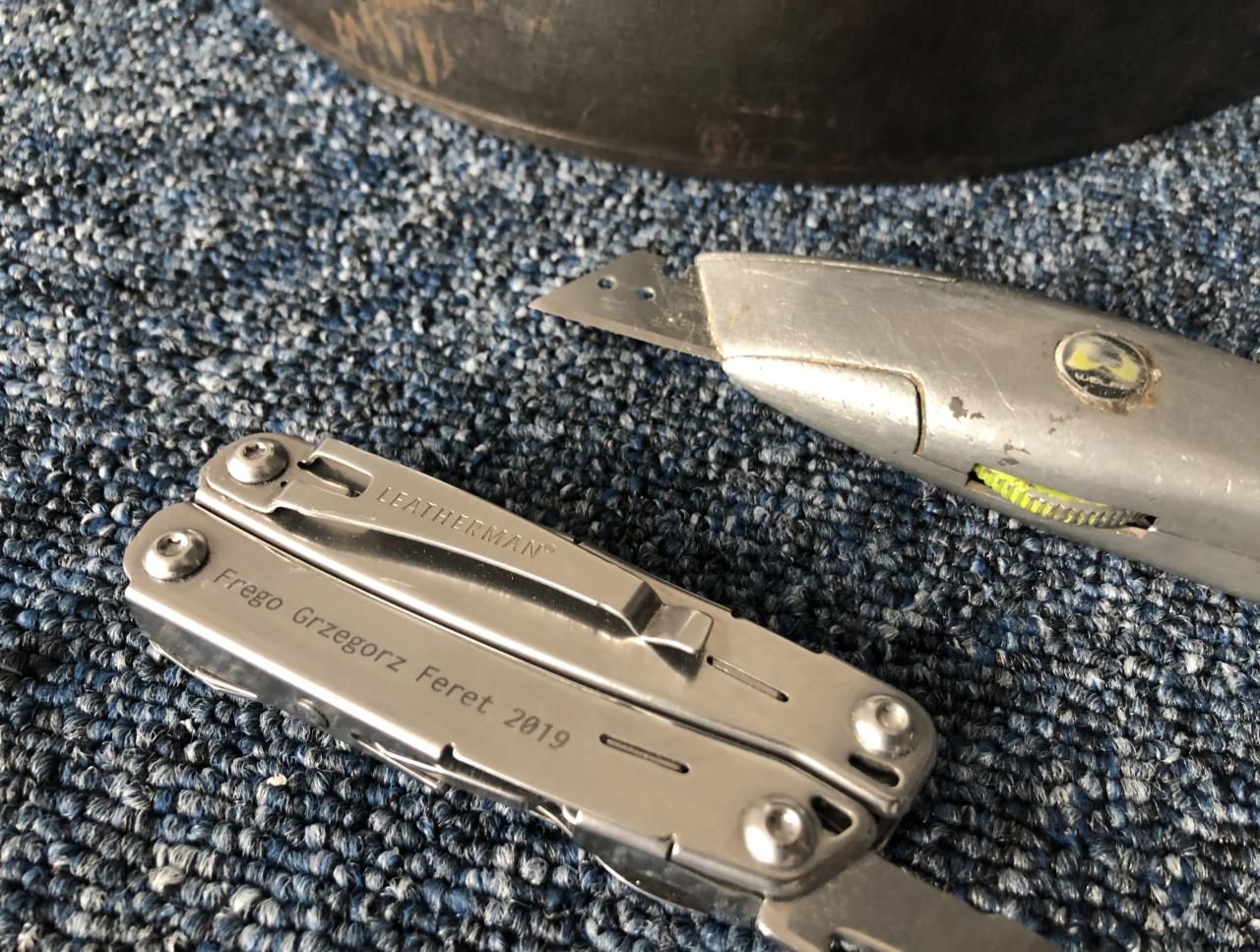 Nóż segmentowy z wysuniętym ostrzem i multitool Leatherman leżą na niebieskiej wykładzinie dywanowej, widoczny fragment czarnego przedmiotu w tle.