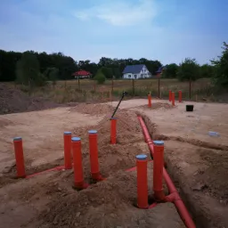Przygotowanie terenu pod budowę domu: widoczne pomarańczowe rury kanalizacyjne z szarymi zaślepkami, wystające z wykopów w ziemi, w tle dwa domy jednorodzinne i linia drzew.