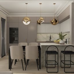 Studio Dobre Miejsce Sylwia Malewicz - Stylowa aranżacja kuchni z wyspą i krzesłami barowymi, oświetlona wiszącymi lampami w kolorze złota. Neutralna kolorystyka ścian i mebli.