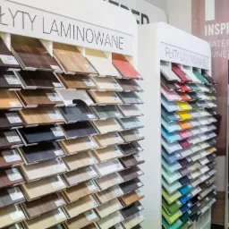 Ekspozycja próbek laminowanych płyt w odcieniach drewna i kolorów, prezentowanych na białych stojakach w sklepie.