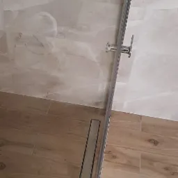 Szklana kabina prysznicowa z chromowanymi okuciami, widoczny odpływ liniowy w podłodze wyłożonej płytkami imitującymi drewno, ściany wykończone płytkami w odcieniach beżu z marmurkowym wzorem.