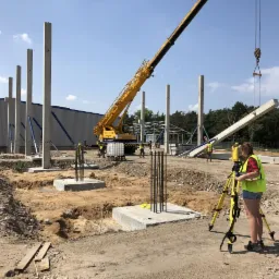 Geodeta w kamizelce mierzy teren pod budowę hali z pomocą niwelatora, w tle dźwig podnosi betonowy element konstrukcyjny.