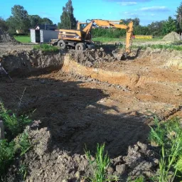 Żółta koparka CAT M315 na skraju wykopu, z widocznym niwelatorem laserowym na statywie, w tle zielona roślinność i budynek gospodarczy.