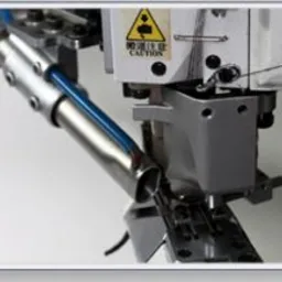 Zbliżenie na głowicę specjalistycznej maszyny szwalniczej z zamontowanym niebieskim przewodem, widoczna tabliczka ostrzegawcza z symbolem trójkąta i wykrzyknika.