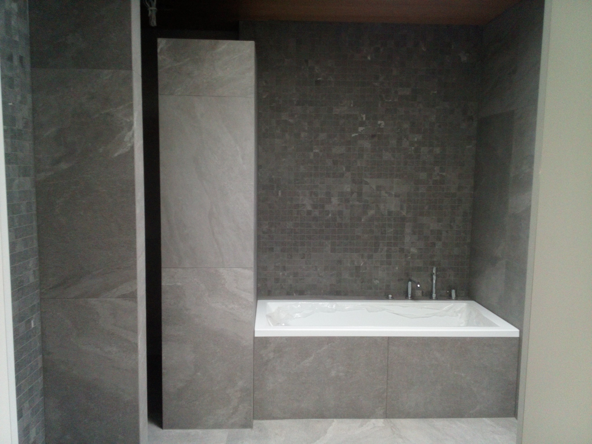 Wanna obudowana szarymi płytami w łazience z mozaiką na ścianie, zabezpieczona folią przed zabrudzeniem podczas prac wykończeniowych.