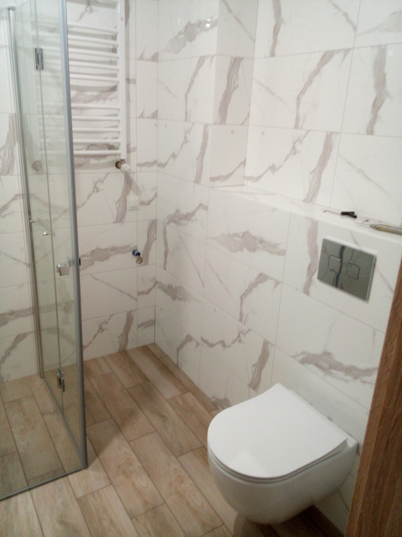 Wykończona łazienka z kabiną prysznicową, grzejnikiem drabinkowym, podwieszaną toaletą i płytkami imitującymi marmur na ścianach oraz panelami podłogowymi.