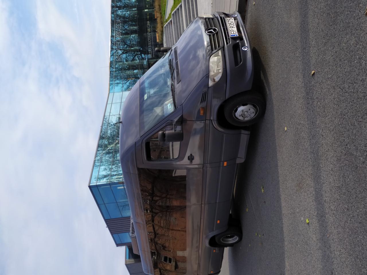 Szary bus Mercedes Sprinter zaparkowany na asfalcie, z budynkiem biblioteki w tle, widoczne odbicie drzew w szybach pojazdu.