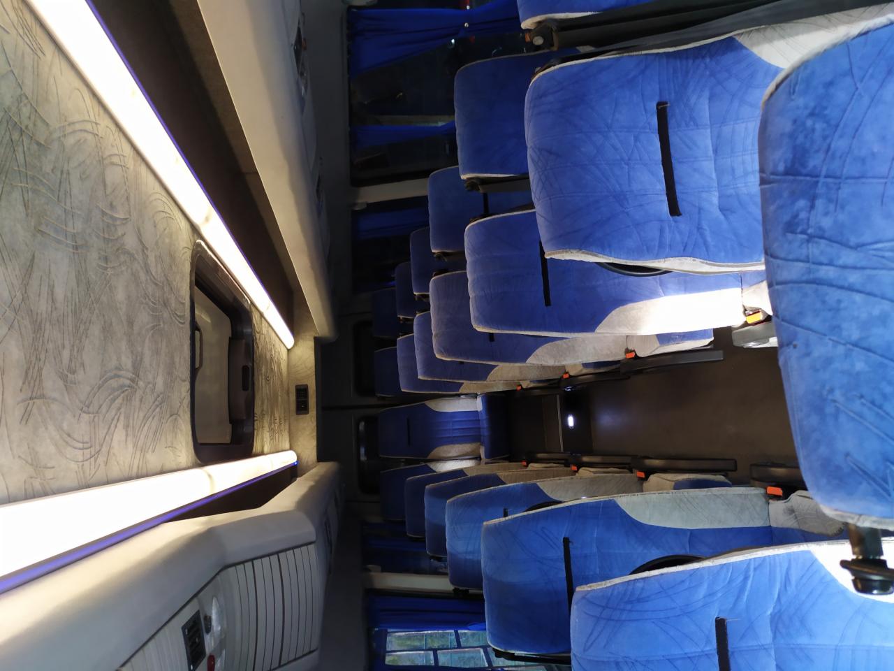 Wnętrze autobusu z rzędami niebieskich, pluszowych siedzeń, oświetlone listwą LED, widok z góry z perspektywy pasażera.